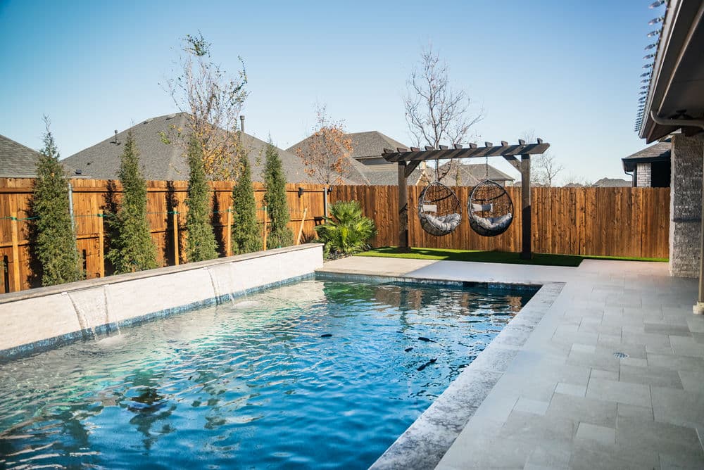 Modern backyard with a swimming pool, hammock chairs, and wooden fence under clear blue sky.