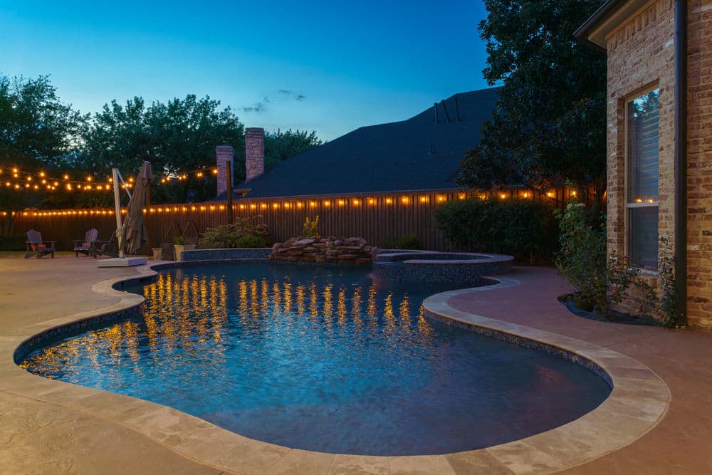 Modern backyard pool at twilight with string lights and lush landscaping.